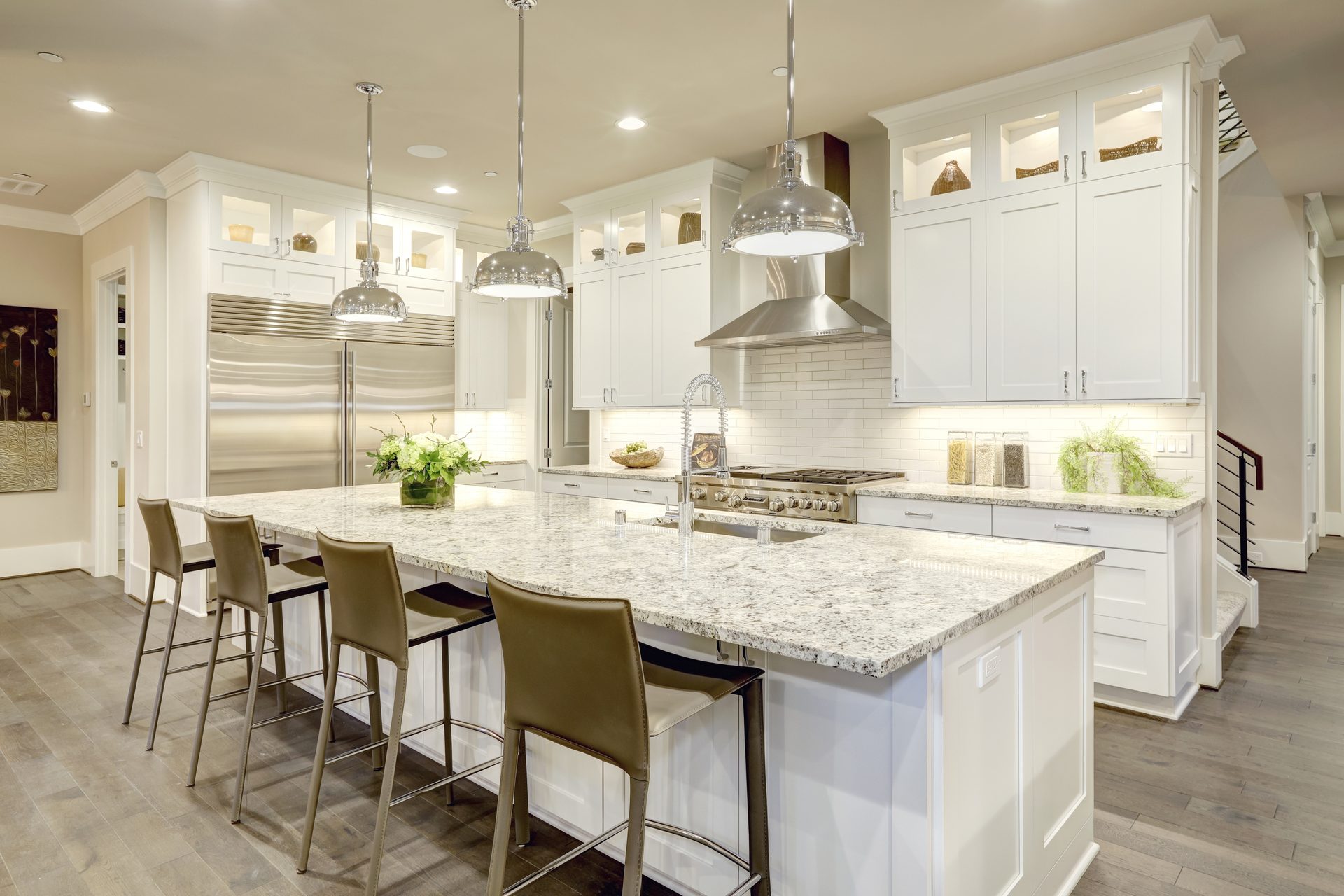 White shaker kitchen with a granite island, tan leather bar stools, chrome pendant lights, and a stainless range hood